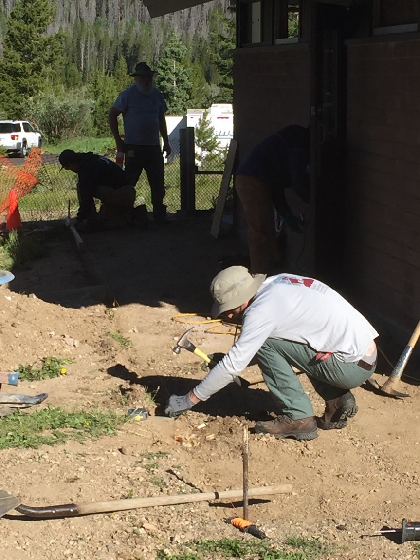 Three people working on a landscaping project outside a brick building, digging and shoveling soil.