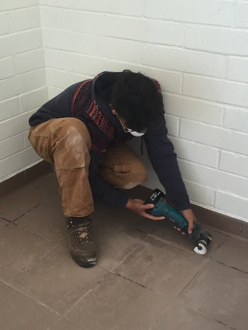 A person in work clothes and glasses uses a power drill on a wall corner.