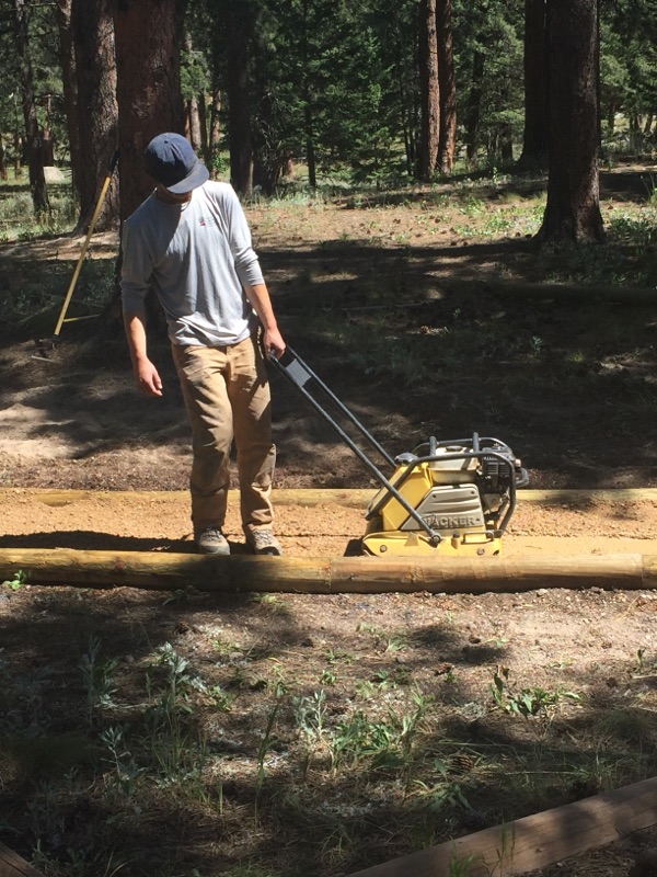 A man in a hat and casual wear using a portable sawmill to cut a log in a forested area.