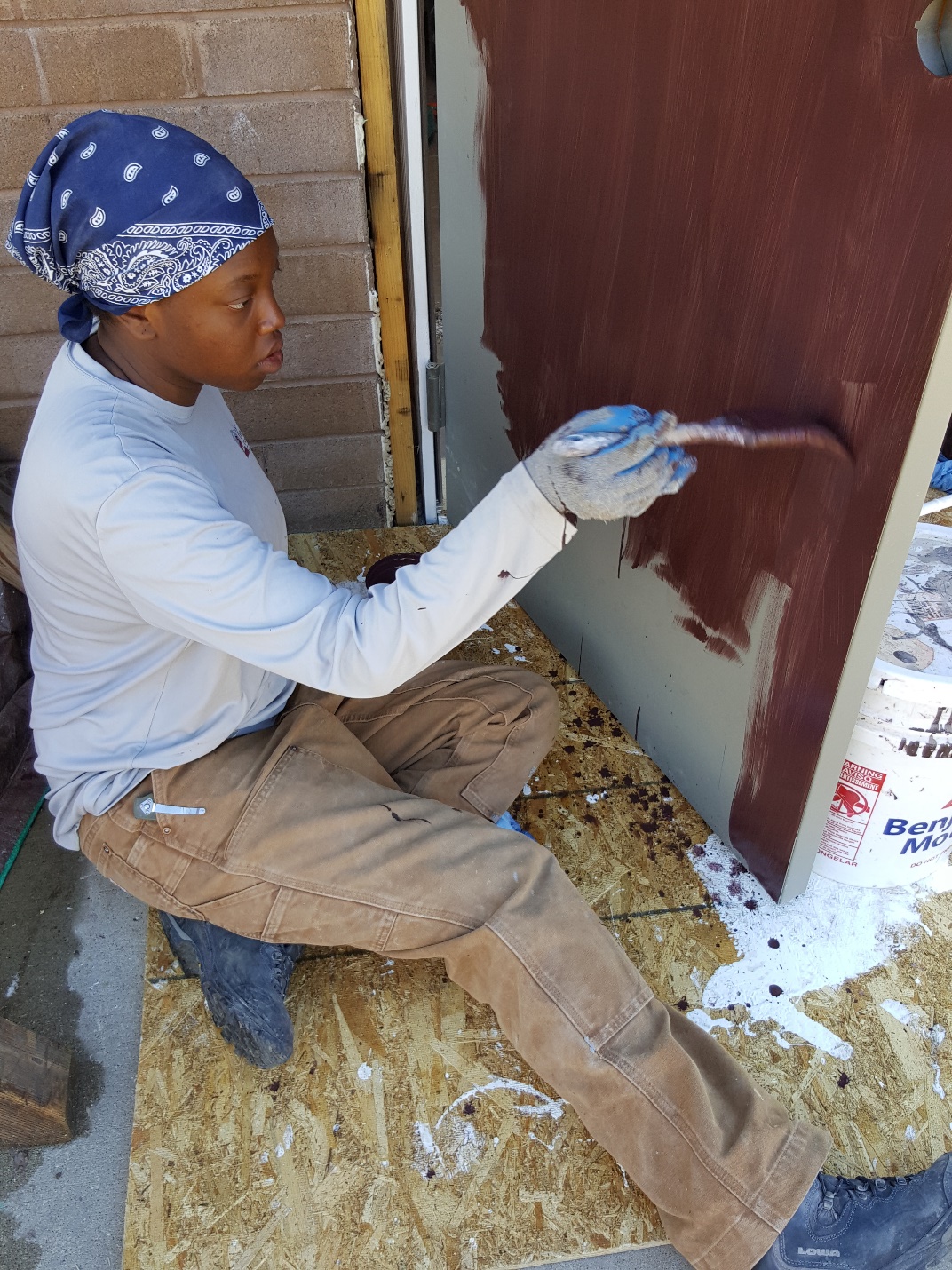 A person in a blue bandana and gloves painting a door brown.