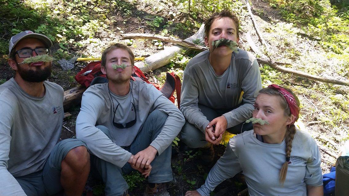 Four people sitting in a forest, each with a leaf held under their nose like a mustache