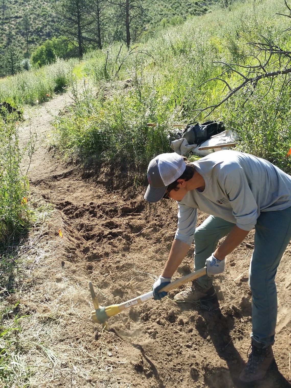 A person using a pickaxe to work on a trail in a sunny, grassy hillside.