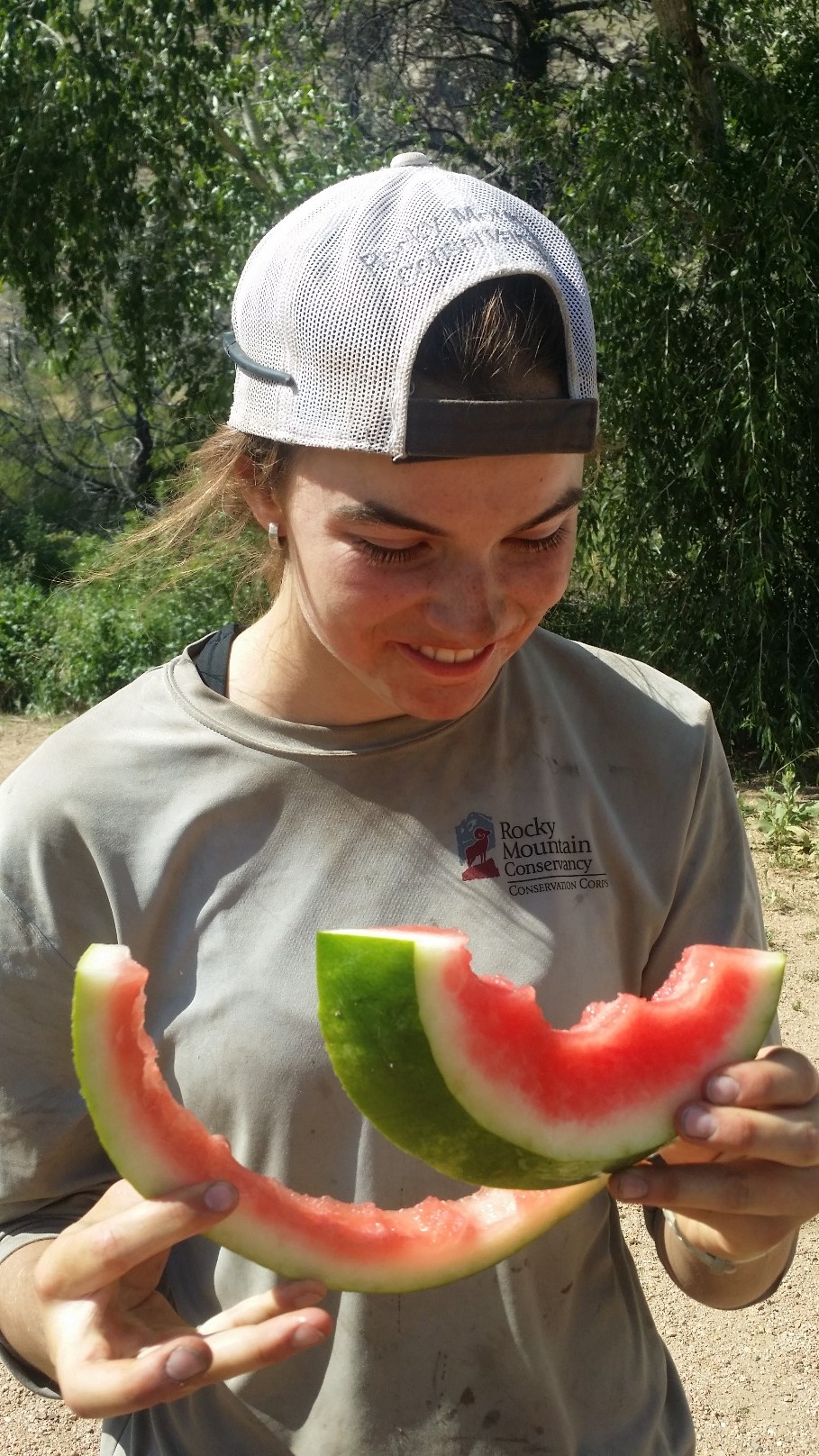 A smiling young woman in a cap and work shirt holds a large slice of watermelon outdoors.