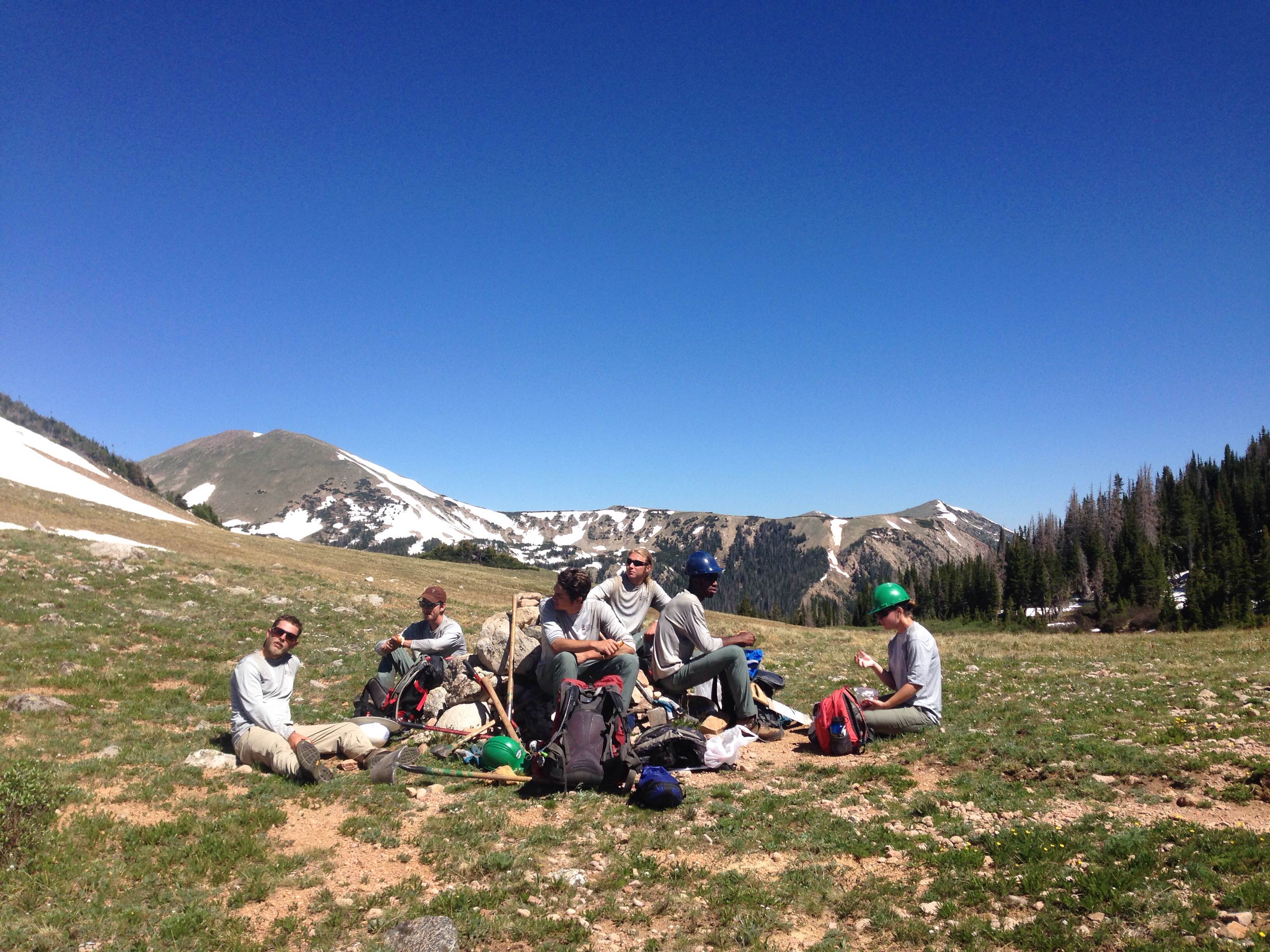 Group of hikers resting with backpacks in a mountainous area with patches of snow.