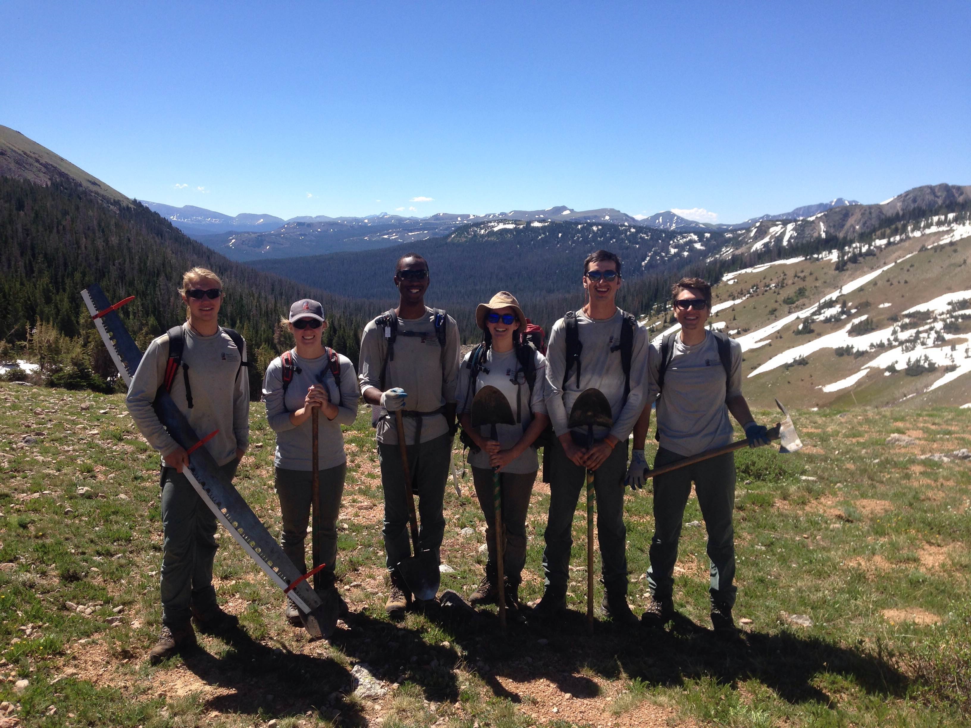 Six volunteers holding tools stand in a mountainous region
