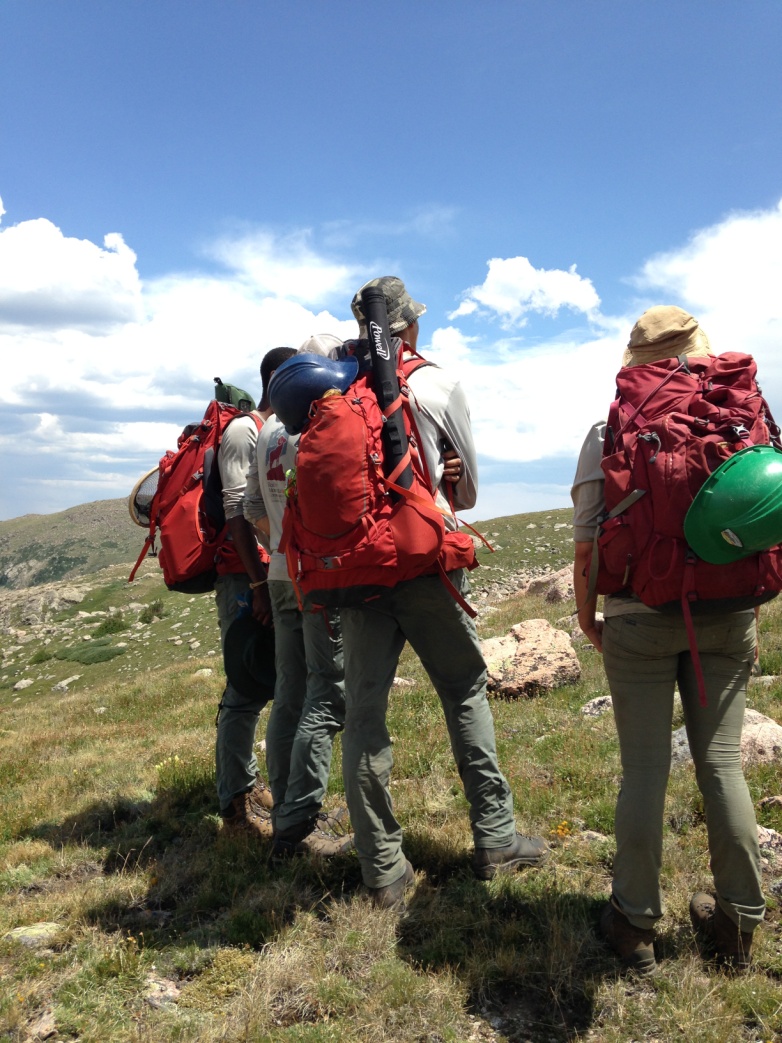 Four hikers with red backpacks observing a scenic mountain landscape under a clear blue sky.