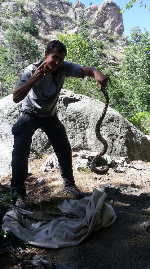 A man smiling and making a peace sign, holding a large snake outdoors near rocks and trees.