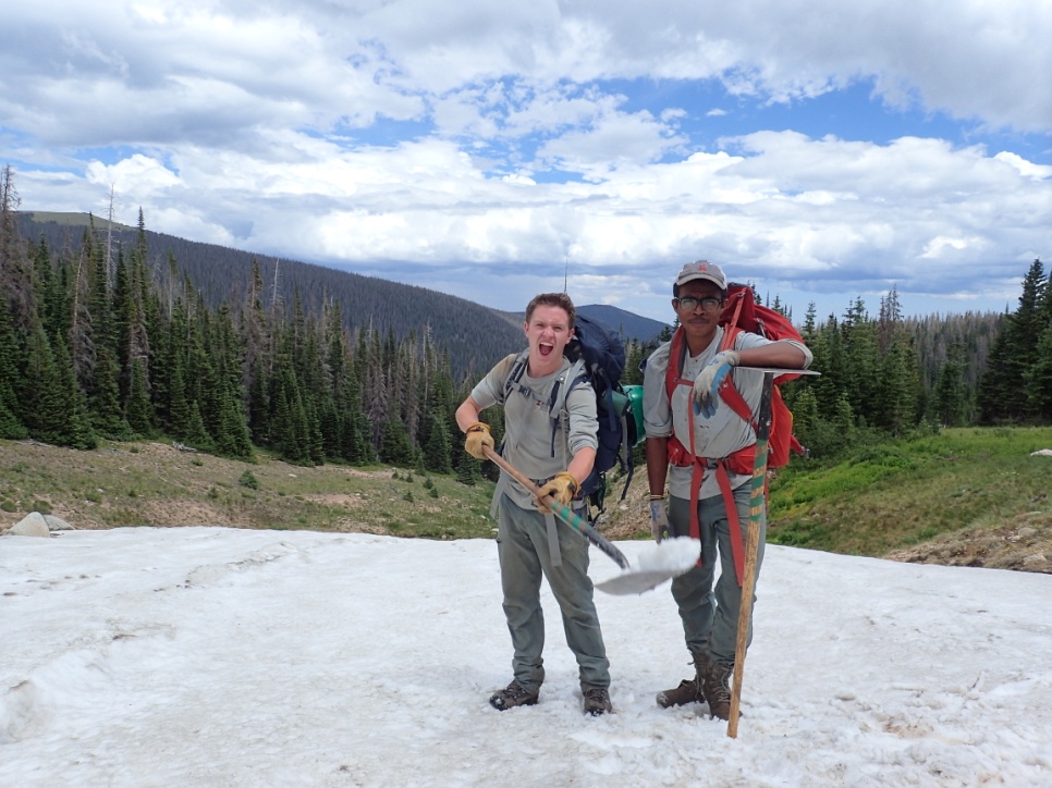 Two hikers smiling while standing on a snowy patch in a forested mountain area