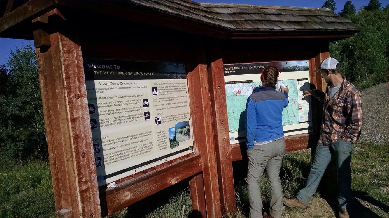 Two people examining a map at a wooden information kiosk in a forested area.