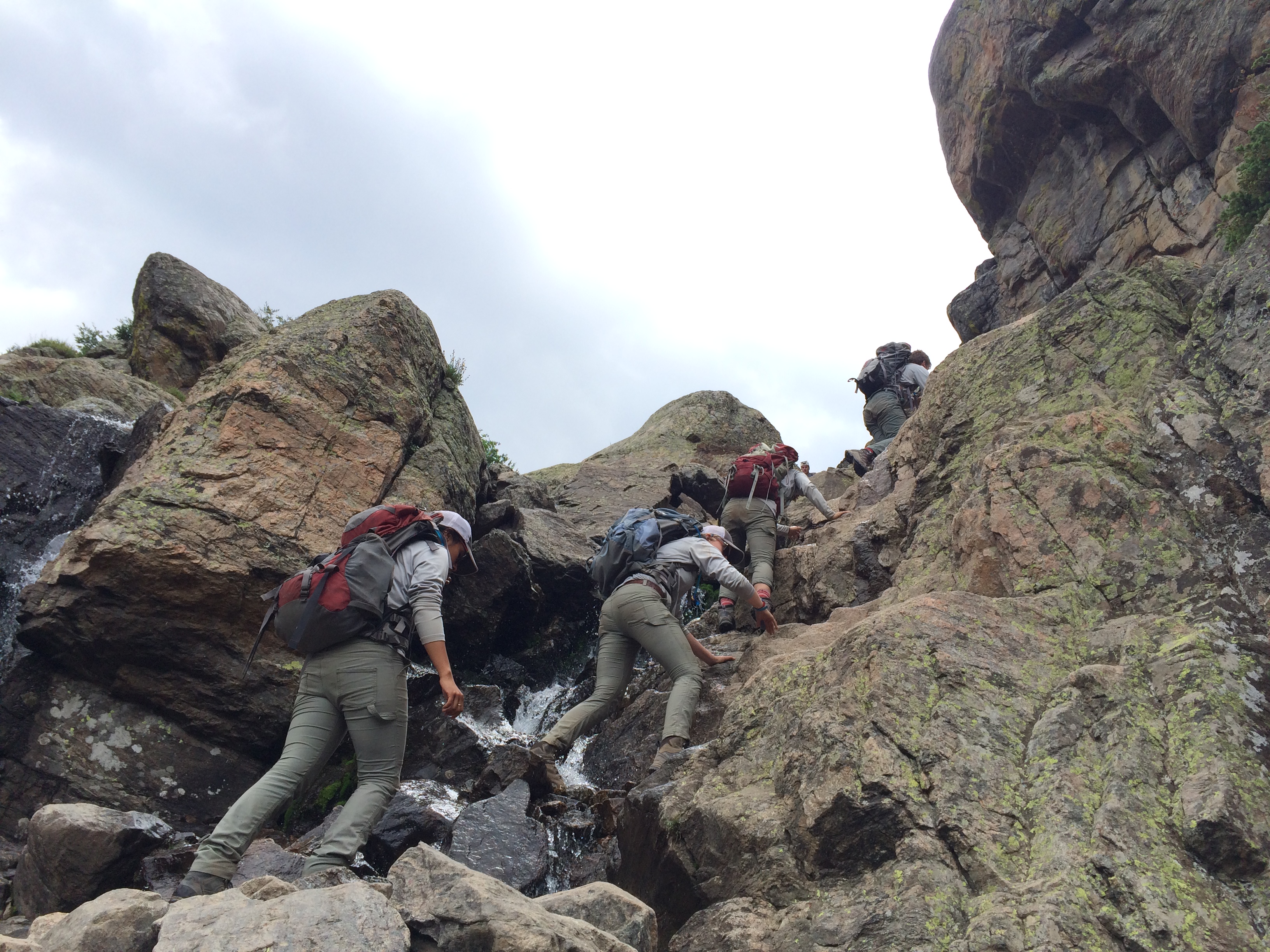 Group of hikers ascending a rocky mountain trail with a small waterfall beside them.