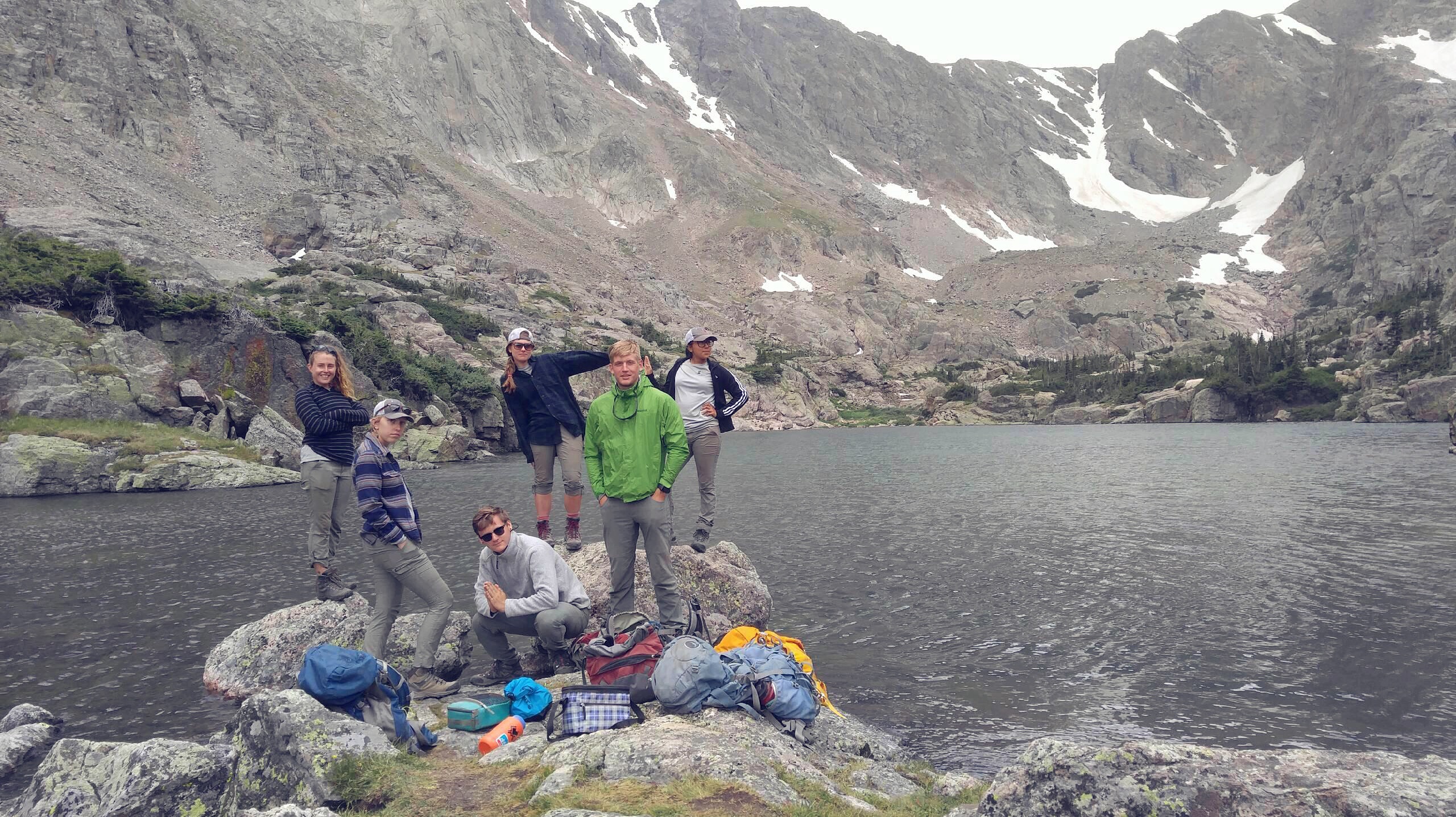 Group of hikers with backpacks posing by a mountain lake with steep rocky slopes in the background.