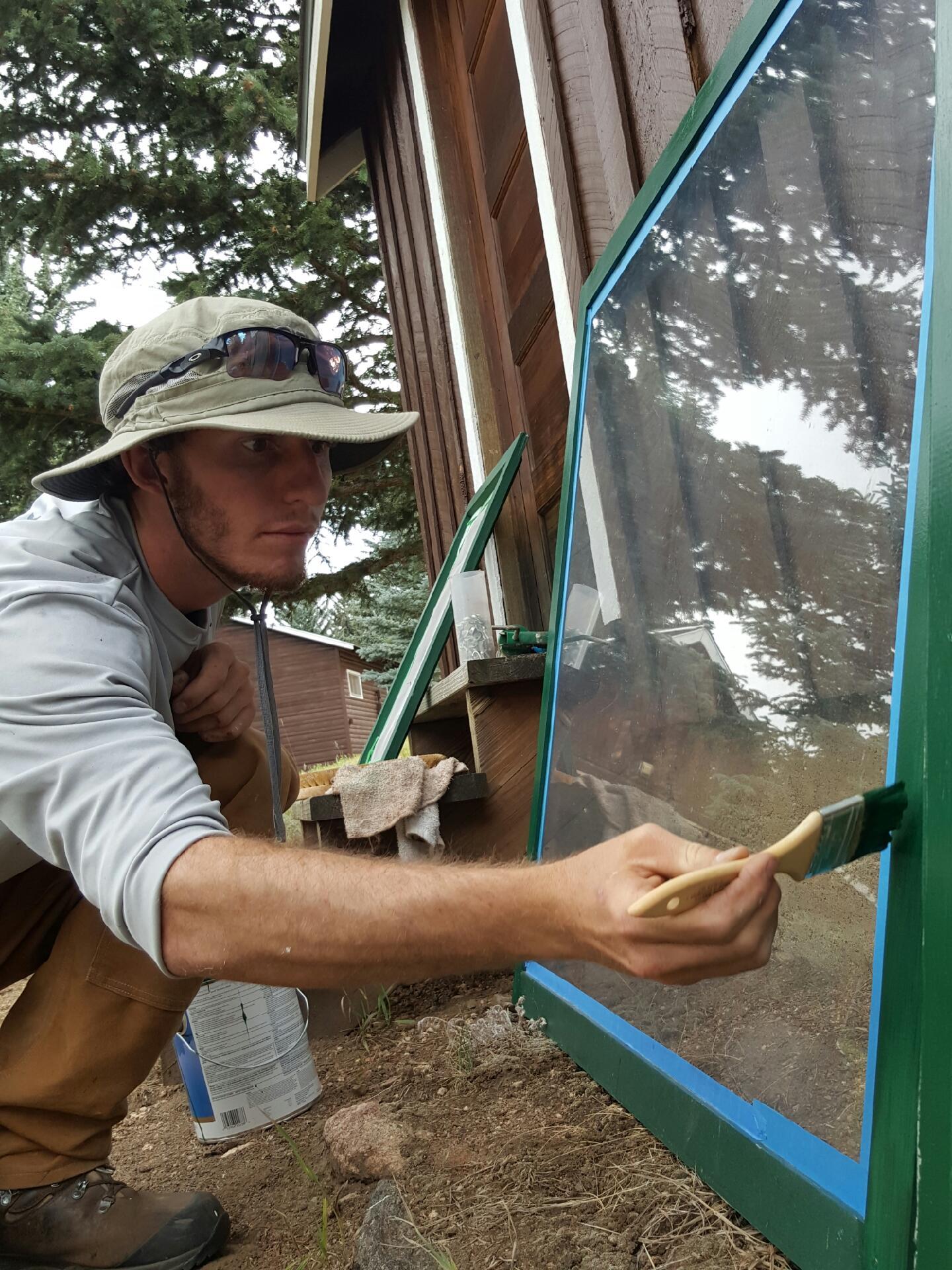 A man in a hat applies paint to a window frame with a brush, focusing intently on his task.