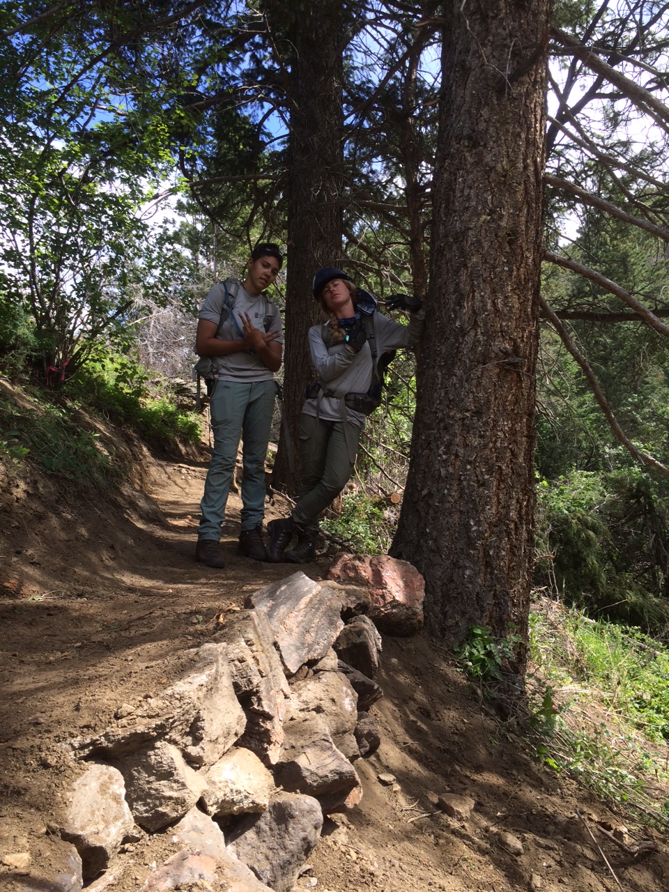 Two hikers leaning against a tree on a dirt trail in a forest smiling at the camera