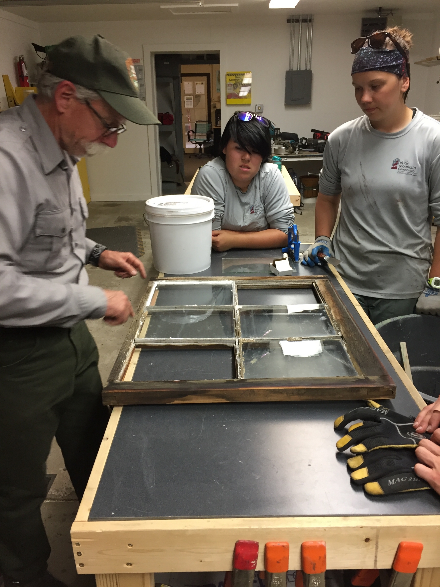 Three people, one older and two younger, examine a wooden window frame on a workshop table