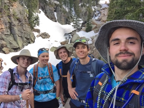 Five young hikers pose for a selfie in front of a snowy mountain pass