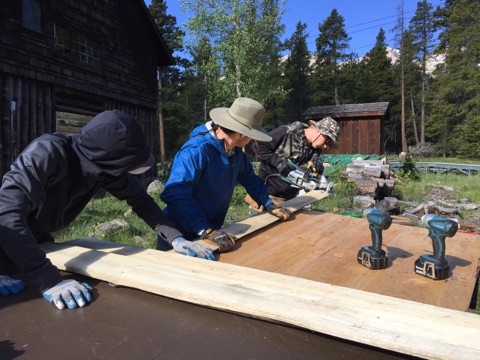 Three people wearing hats and gloves work together to drill into a large wooden plank