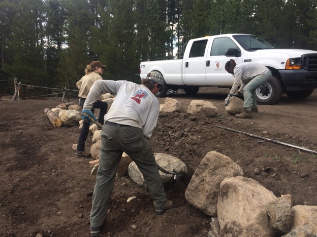 Three people using tools to rearrange large stones in a forest clearing