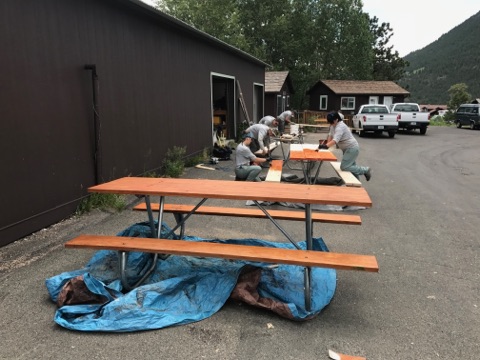 Three people working on sanding and painting a wooden picnic table outside a building