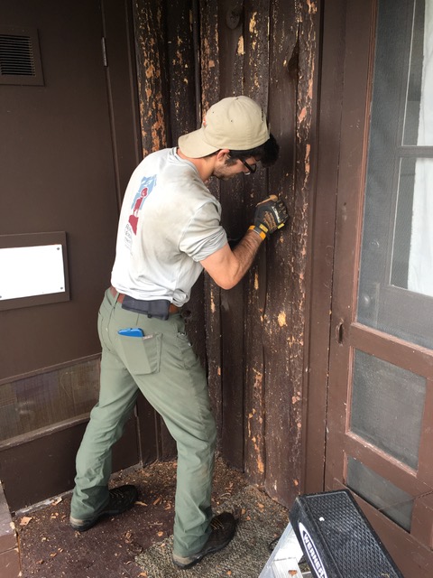 A worker in a cap, mask, and uniform uses a power drill on a weathered wooden door.