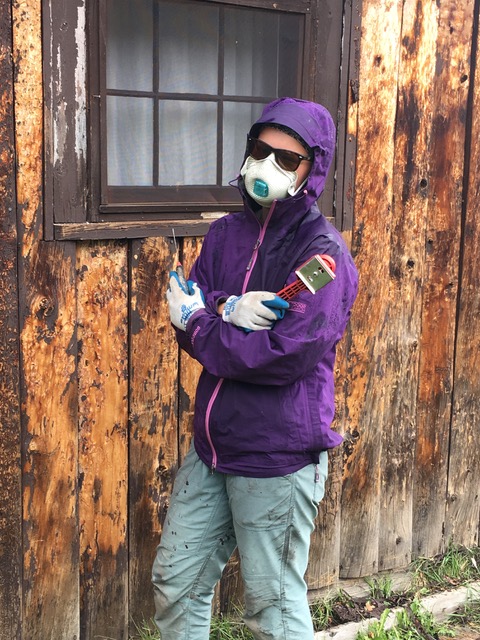 Person in a purple jacket, mask, and gloves standing against a rustic wooden wall with a window, holding paint supplies.