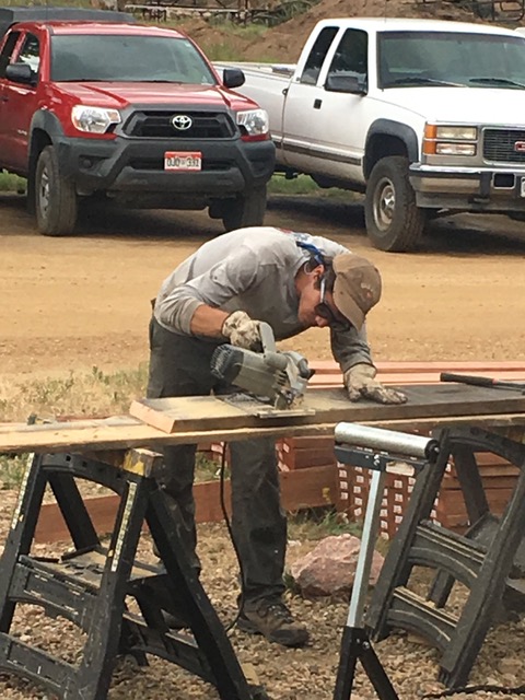 A construction worker in protective gear sawing wood on sawhorses in a dirt-covered area with trucks parked in the background.