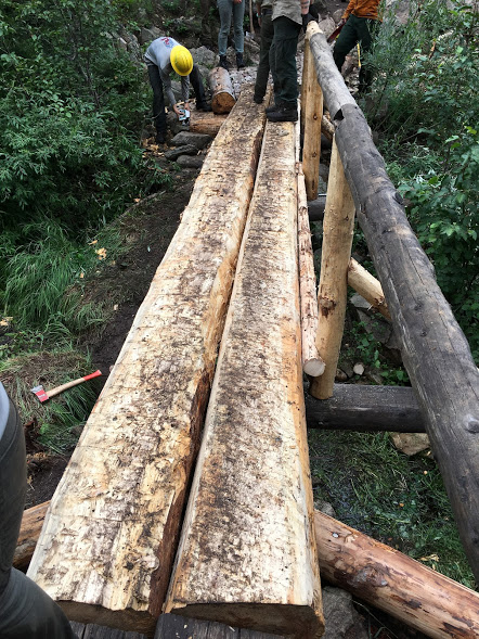 Group of people working on constructing a wooden bridge in a forested area, with tools visible around.