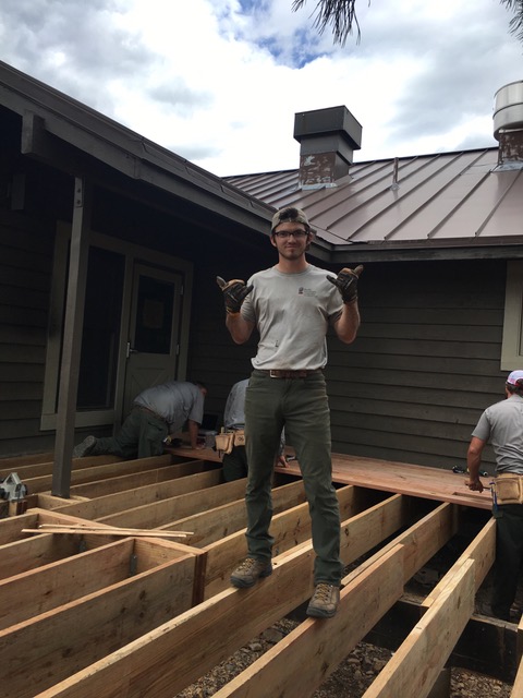 A man in work clothes stands proudly on wooden framing at a construction site, holding gloves, with a house and trees in the background.