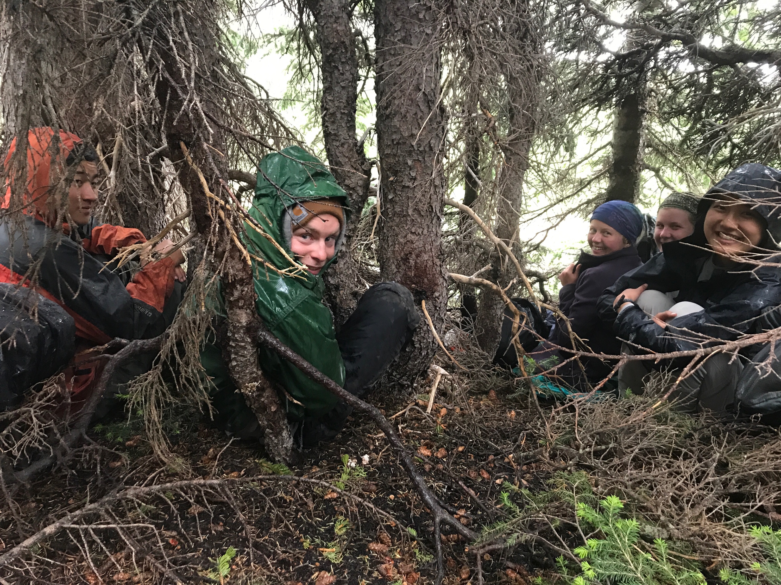 Five people wearing rain gear sheltering under trees, smiling and huddled together on wet ground.
