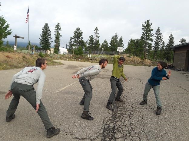 Four people wearing similar outfits playfully imitate a ninja fight stance in a parking lot, with trees and a flagpole in the background.