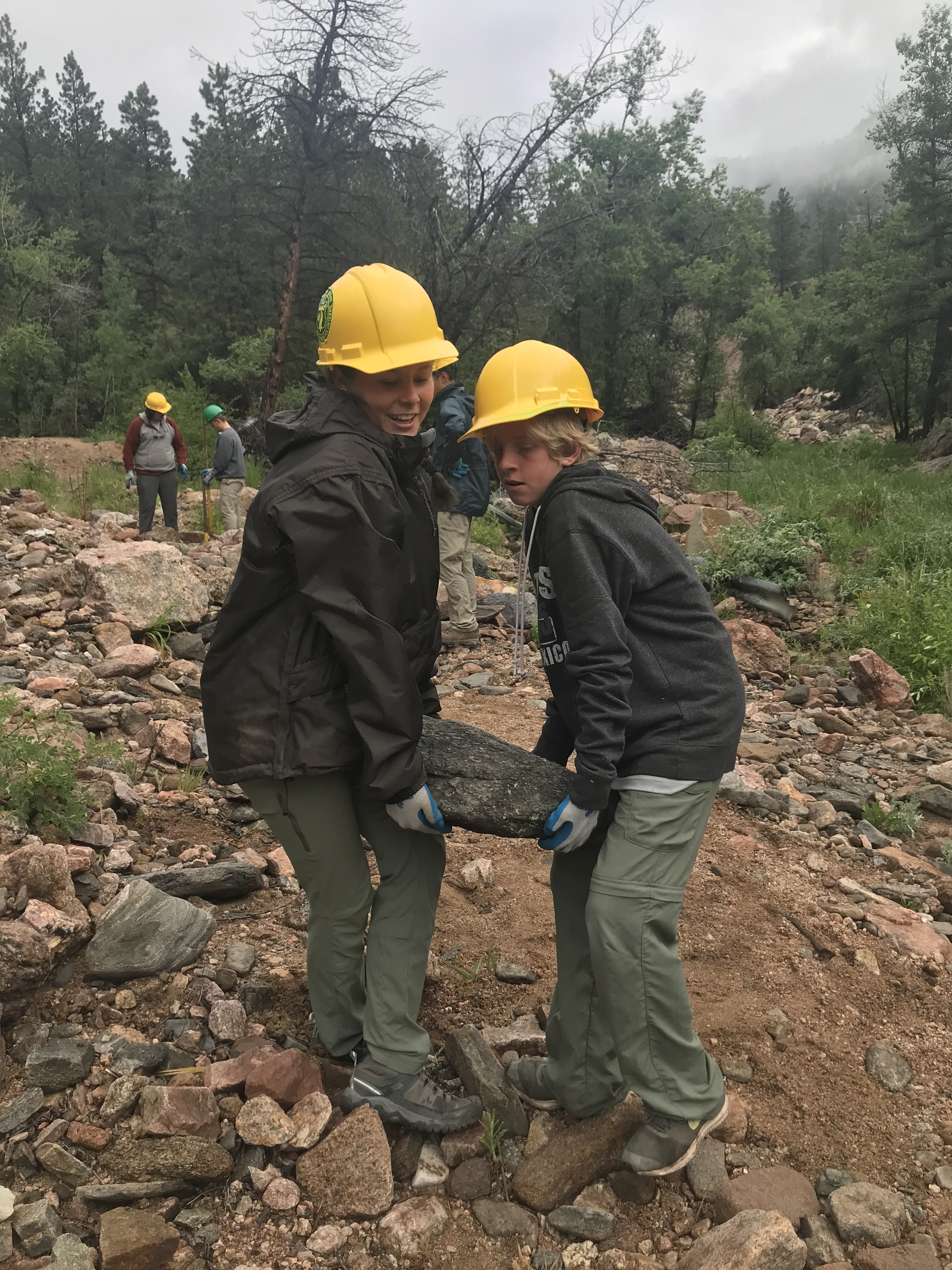 Two children in hard hats and jackets carry a large rock together