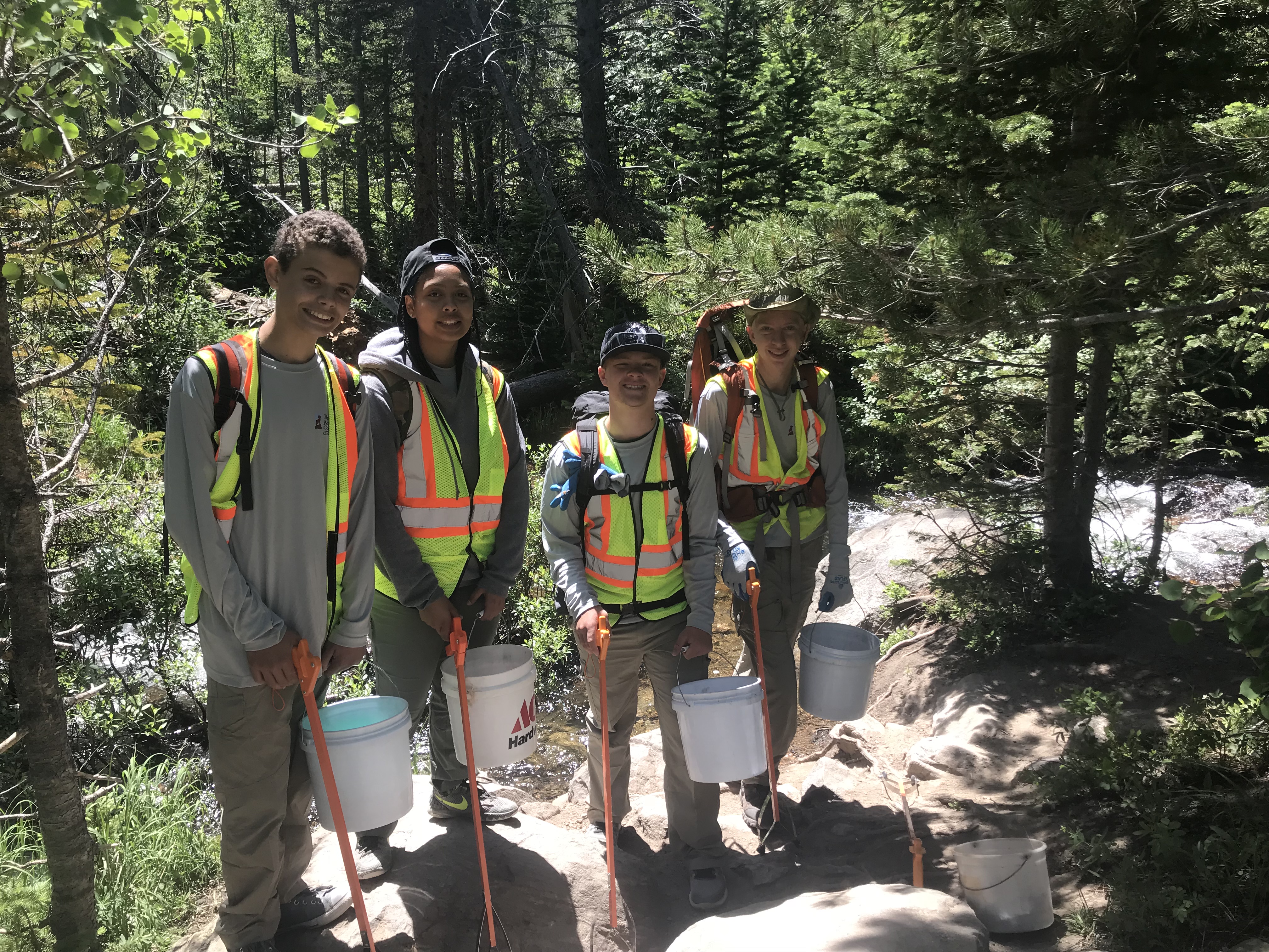 Four trail workers in high visibility vests hold tools and buckets on a forest path next to a river.