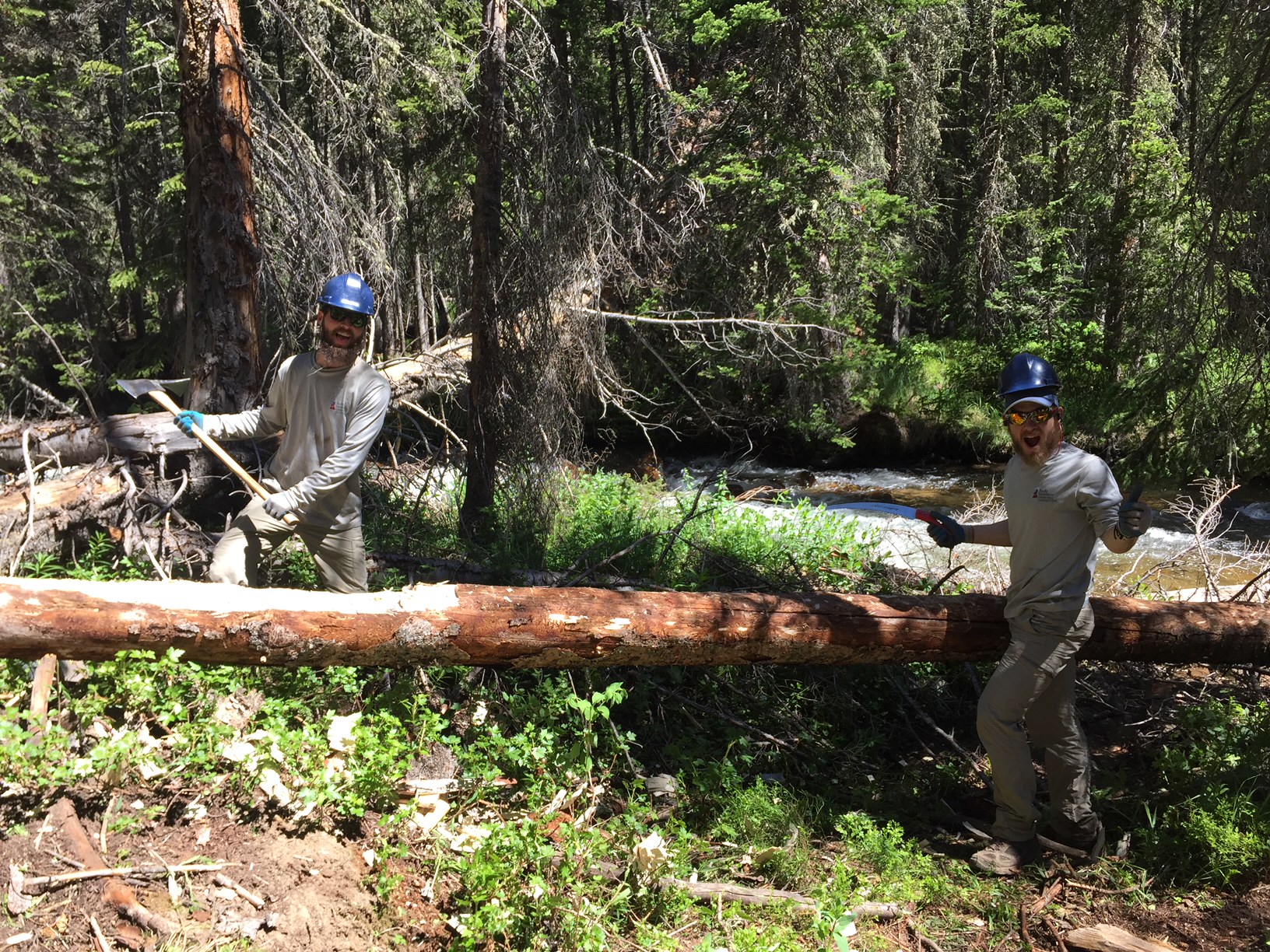 Two people in helmets and gloves carry a large log in a forest near a stream.