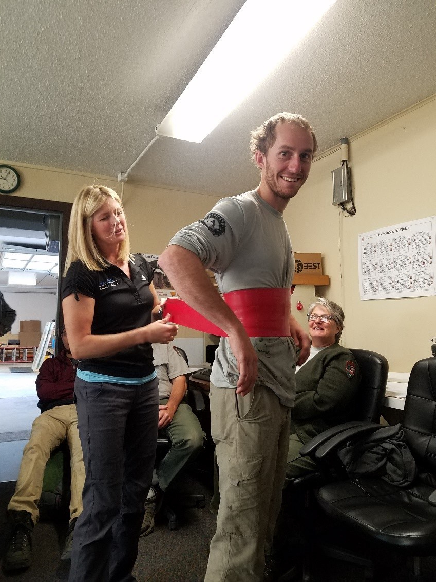 A woman assists a smiling man by adjusting a red back brace around his waist