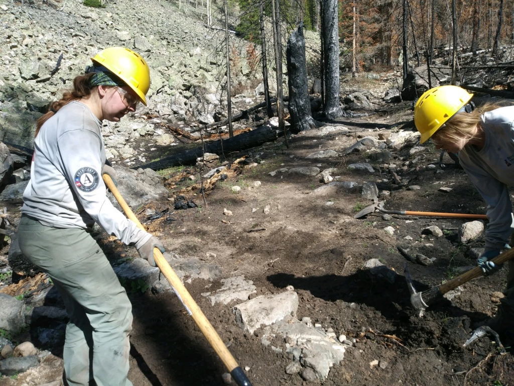 Two women working with a shovel in a wooded area.