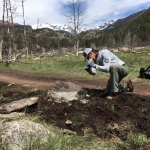 A man kneeling on a dirt trail with mountains in the background.