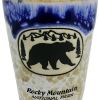Blue Bear Shot glass with a bear silhouette, mountain, and tree design. Text reads "Rocky Mountain National Park" with a two-tone blue upper section and textured earthy lower section.