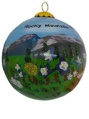 The Ornament RMNP Glass is a round piece featuring a painted landscape of the Rocky Mountains, showcasing mountains, a green field, and colorful flowers.