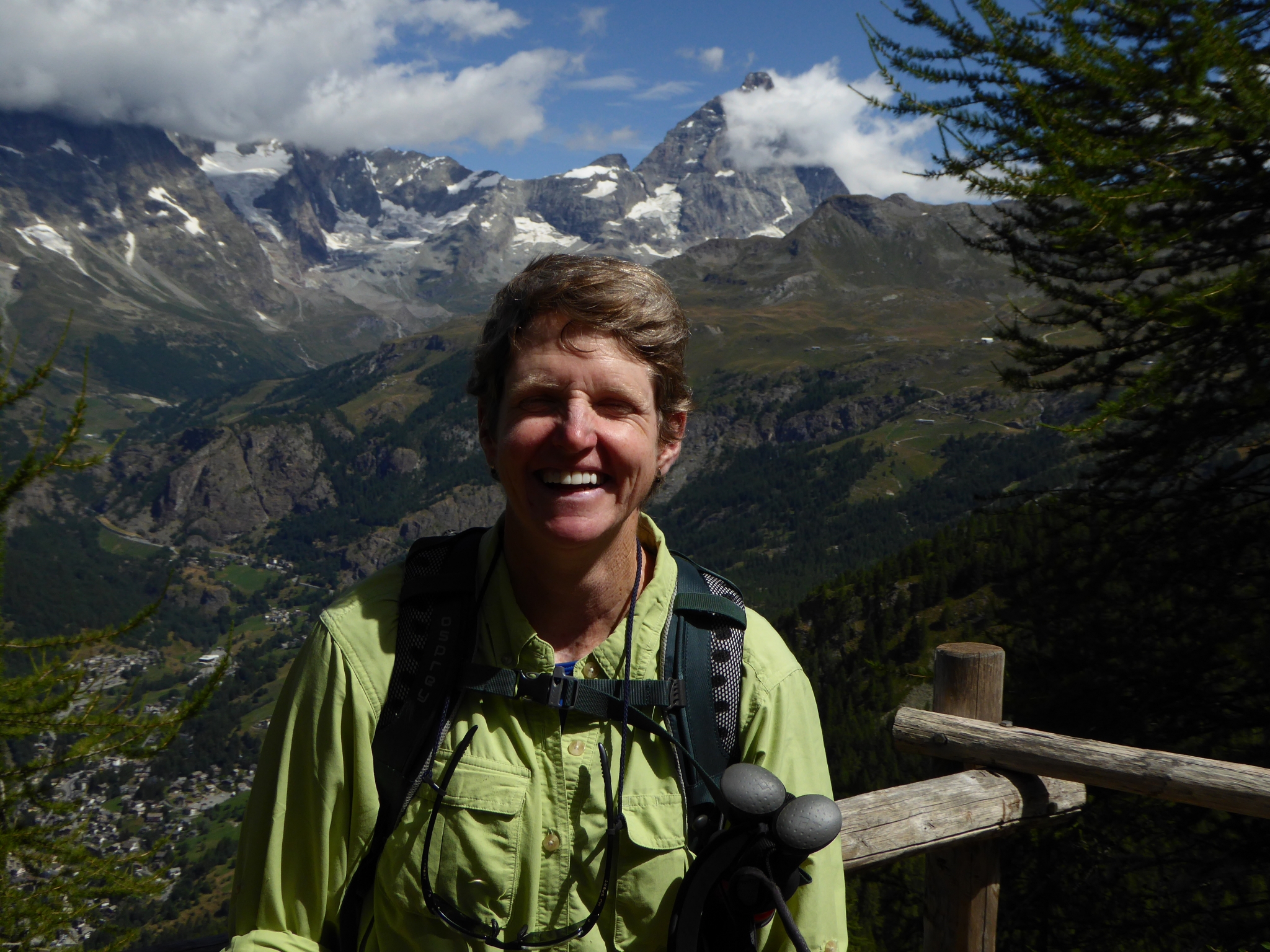A smiling person with a backpack standing in front of a mountainous landscape.
