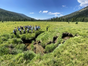 A group of people stand in a grassy meadow near a small stream, surrounded by green hills and trees under a clear blue sky.