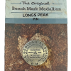 A "Longs Peak Summit Marker" pin in packaging, showing an elevation of 14,255 feet and labeled as "The Original Benchmark Medallion" by Geo Situ.