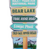 The "Magnet - RMNP Sign" features colorful arrows pointing to Bear Lake, Trail Ridge Road, Longs Peak, and Old Fall River Road beneath "Rocky Mountain National Park," with snowy mountains and a lake in the background.