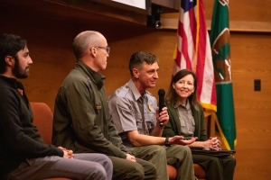 Four people seated on a panel, with one person holding a microphone. U.S. and organizational flags are visible in the background.