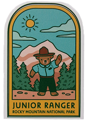 Illustration of a bear in ranger outfit with mountains, trees, and a sun. Text: "Junior Ranger Rocky Mountain National Park." Product: Sticker - RMNP Junior Ranger Bear.