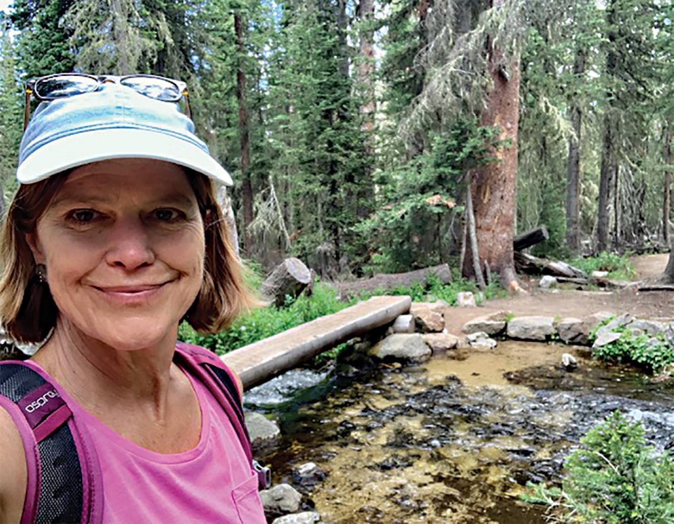 Person in a pink shirt and cap stands near a stream in a forested area, with trees and rocks visible in the background.