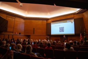 Audience in a wood-paneled lecture hall listens to a presentation with a slide displaying a QR code and text about mountains.