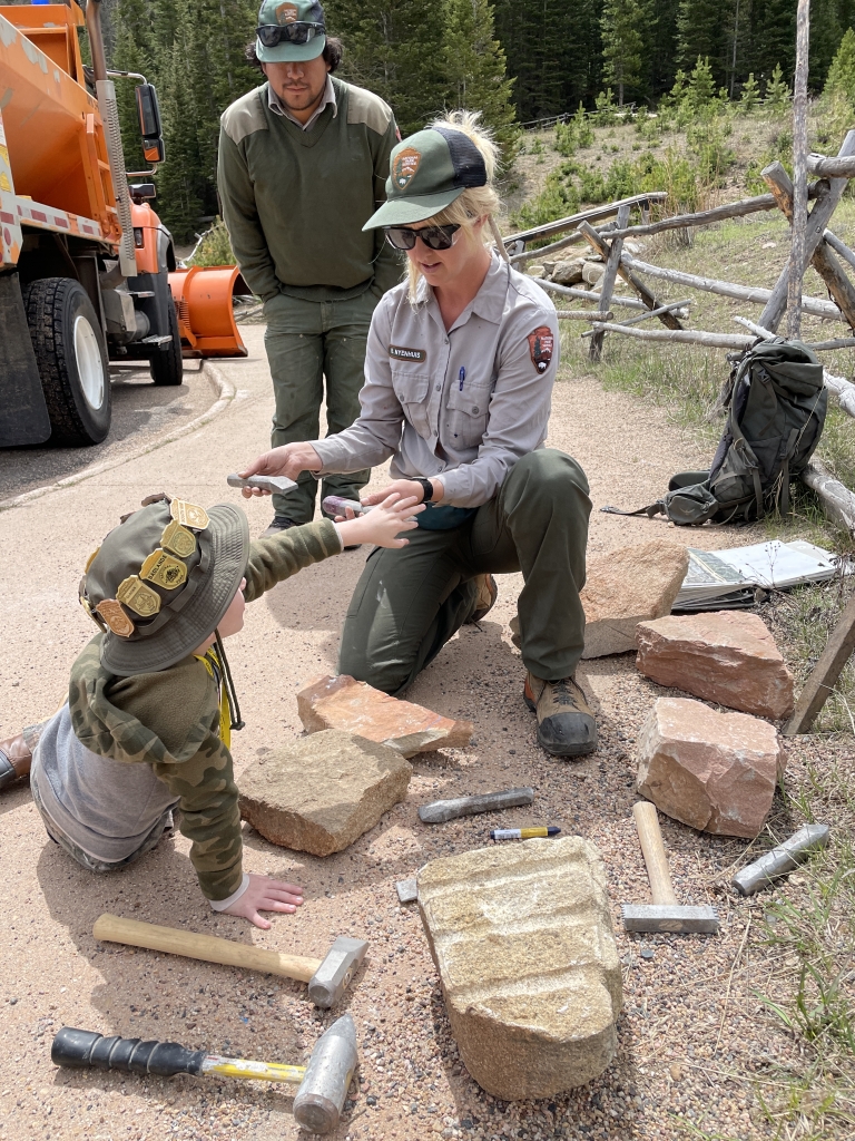 A park ranger kneels, showing a young child an object in her hand. Another adult stands beside them. Various tools and rocks are scattered nearby.