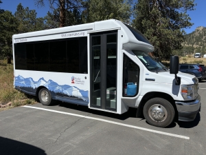A white shuttle bus with mountain graphics and "Rocky Mountain Conservancy" branding is parked in a lot near trees and mountains.