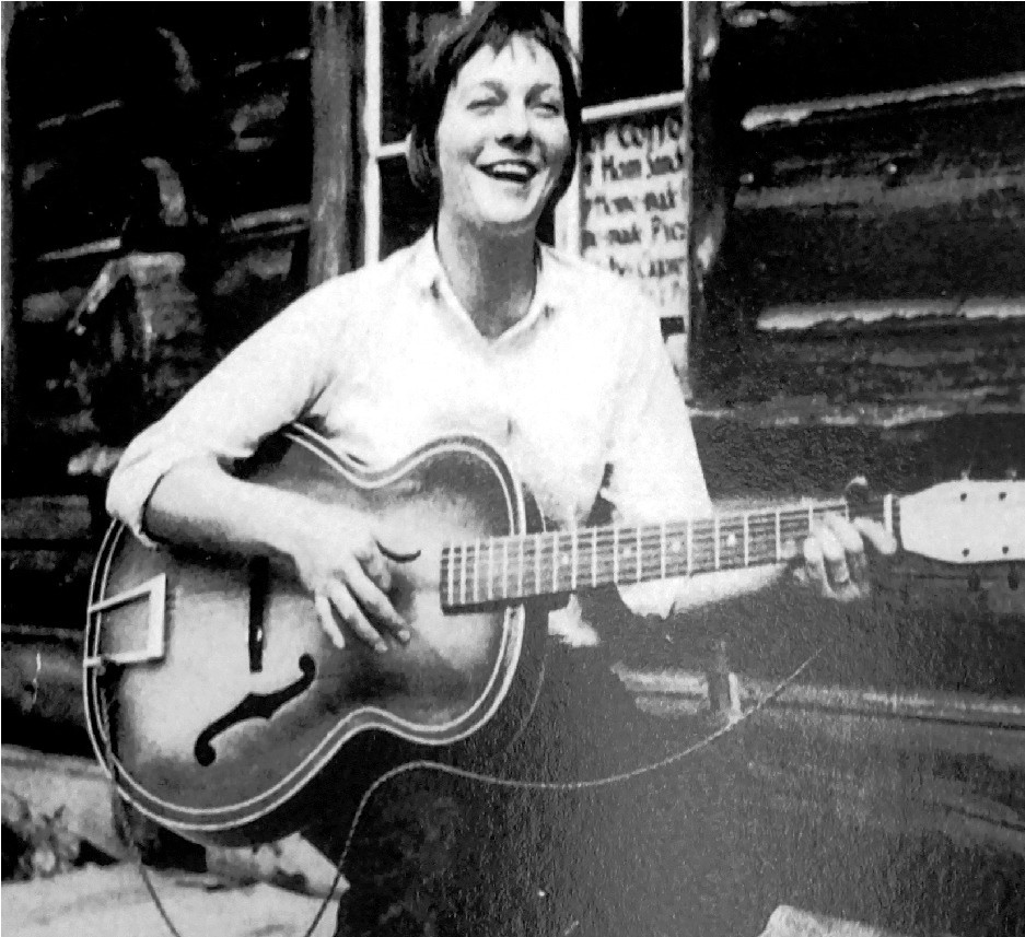 A person sits outside in front of a wooden building, smiling and playing an acoustic guitar.
