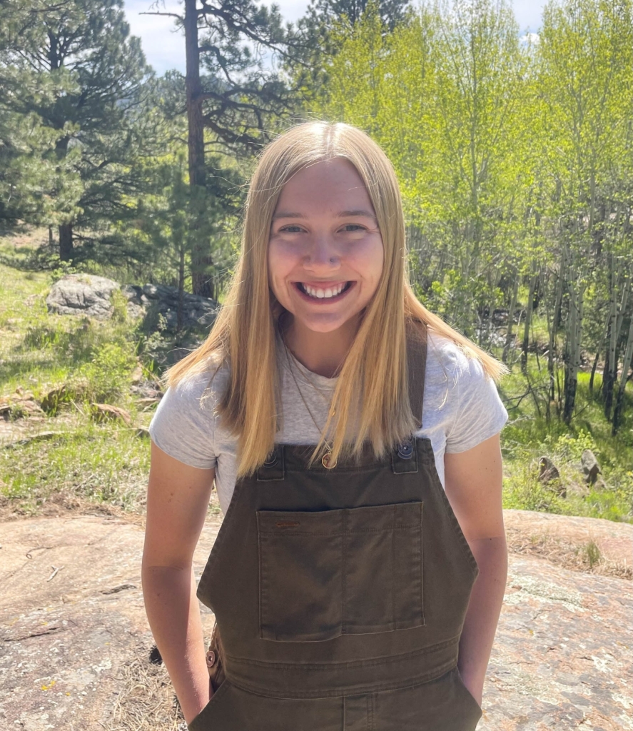 A person with straight blonde hair, wearing a white shirt and olive green overalls, stands outdoors on a sunny day with trees and rocks in the background.