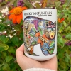 A hand holds the Mug - RMNP Bear Collage, a white cup with a colorful bear illustration featuring animals and plants; orange flowers and green leaves are in the background.